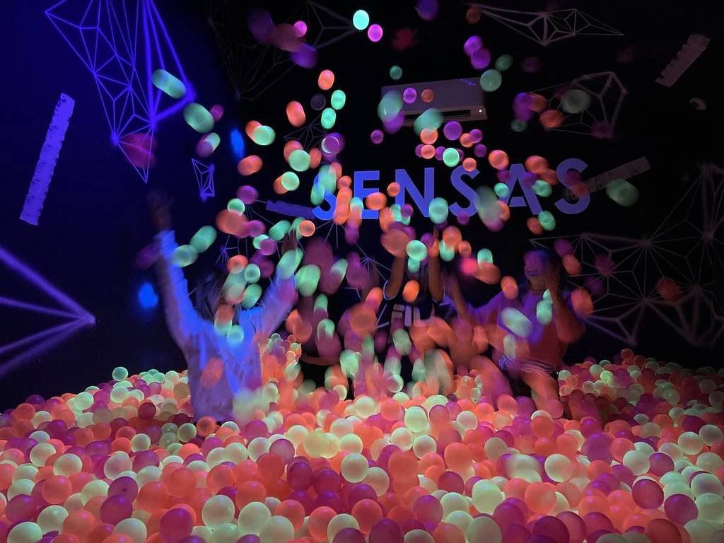 A group of people are playing in a ball pit at SENSAS Melbourne. It is dark and the balls appear to be glowing.